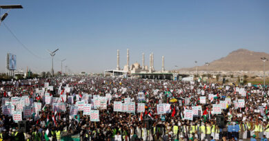 A large crowd, many holding up signs in Arabic, march on a street. There is a mosque in the background and a hill.