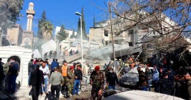 A crowd of people, some in camouflage clothing, gathers outside by a collapsed building.