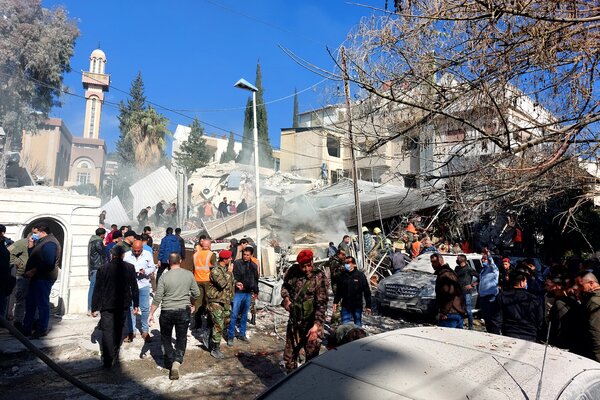 A crowd of people, some in camouflage clothing, gathers outside by a collapsed building.