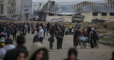 People walk on a road carrying bags, bottles of water and other items. There are tanks in the background and damaged buildings.